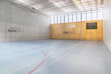 One of the three courts in the Sport-Toto sports hall. One wall is in concrete, one in light-wood panels and one is the curtain separating the court with the next. We don't see the fourth side. The floor is light blue and has many lines.