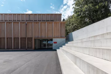 The Training hall from the outside. It is covered in wood-panels. On the side, we see large stairs made of concrete.